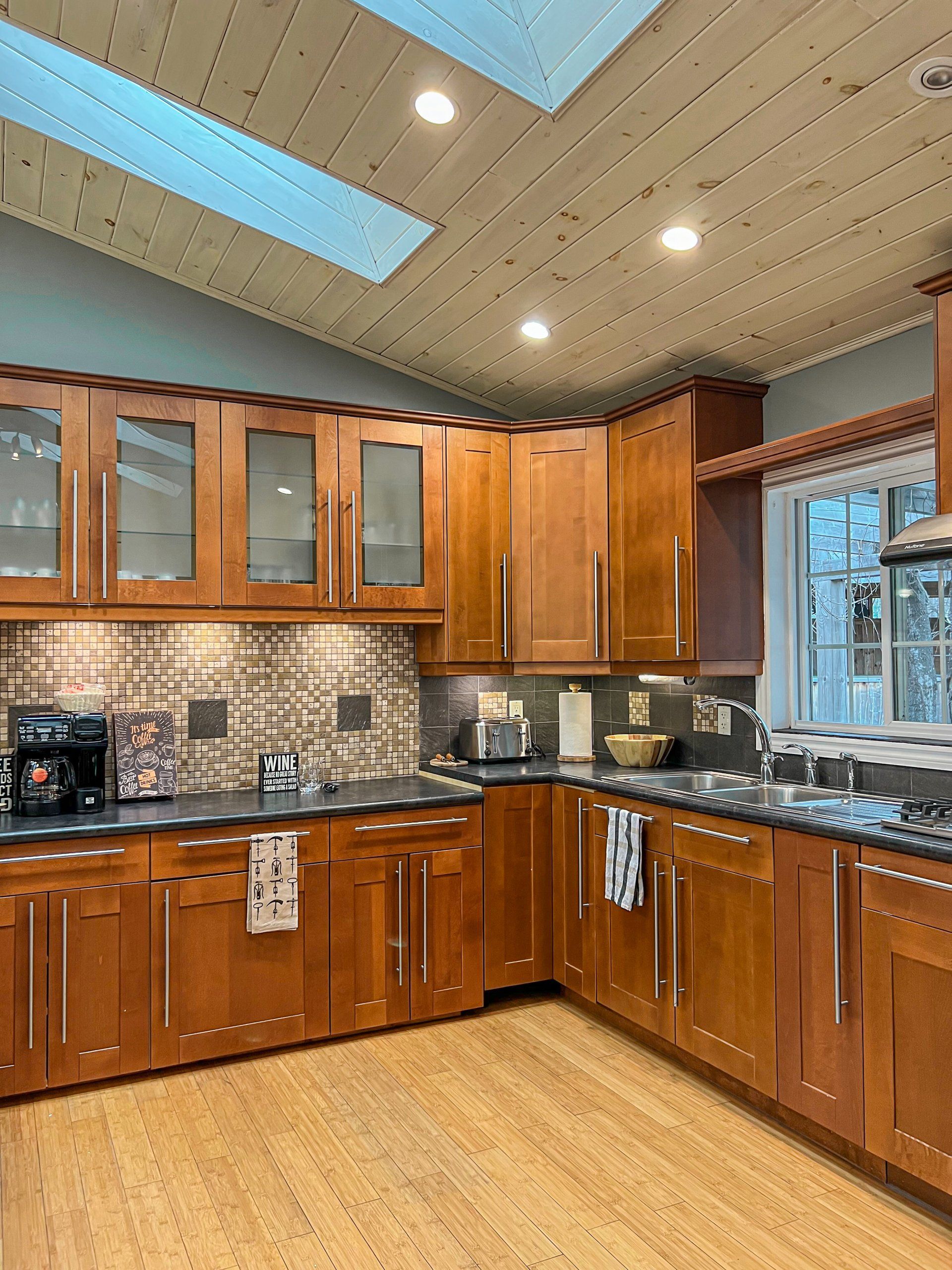 Modern kitchen with wood cabinets, tile backsplash, black countertops, and skylights over hardwood floor