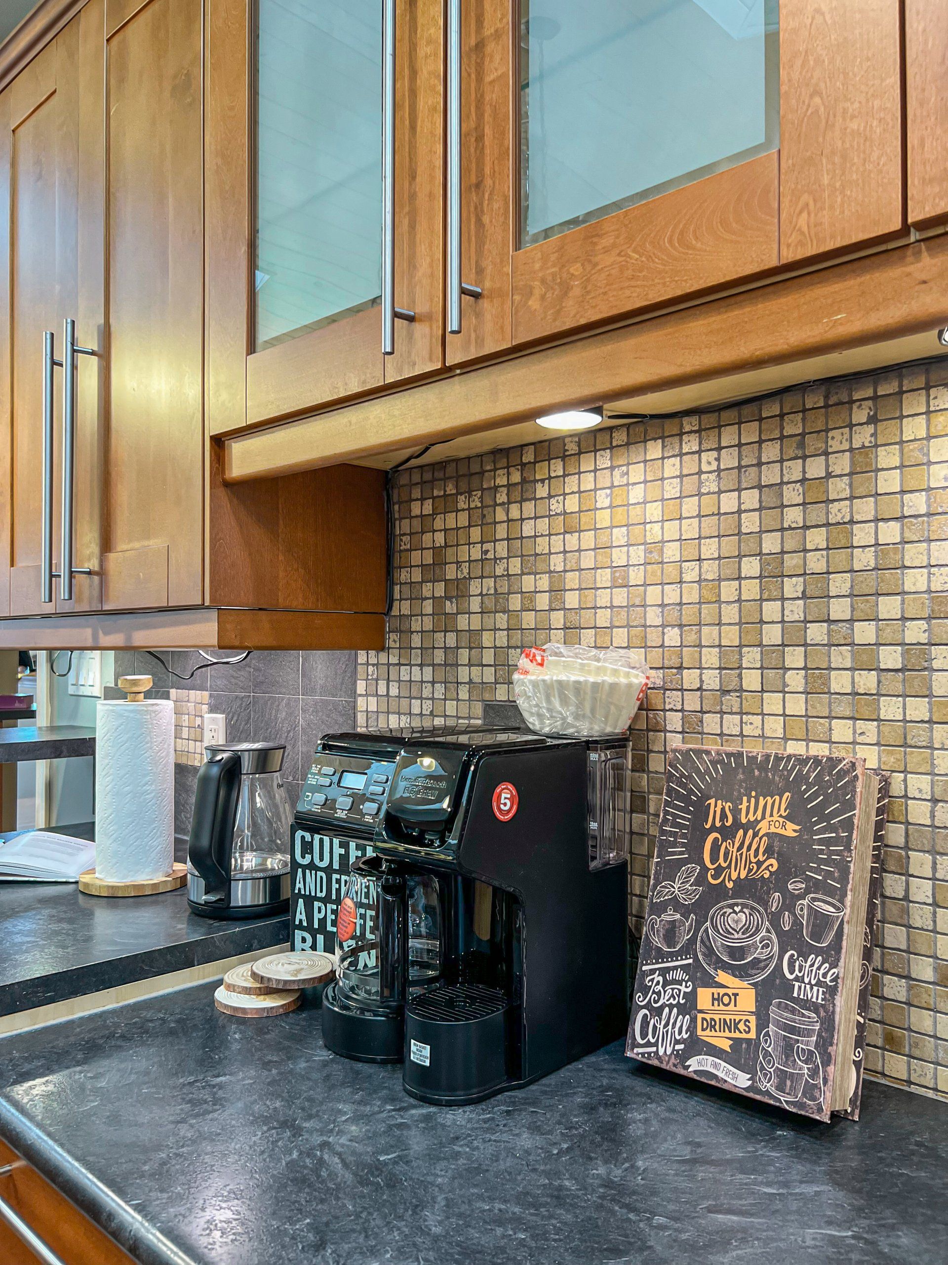 Kitchen countertop with coffee maker, mugs, and a decorative sign beneath wooden cabinets.