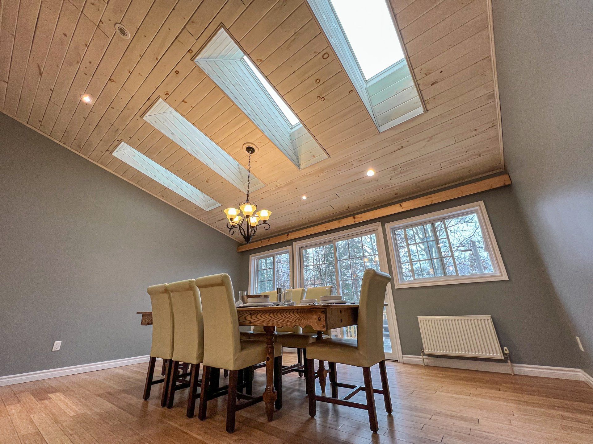 Modern dining room with a wooden table, beige chairs, skylights, chandelier, and gray walls.