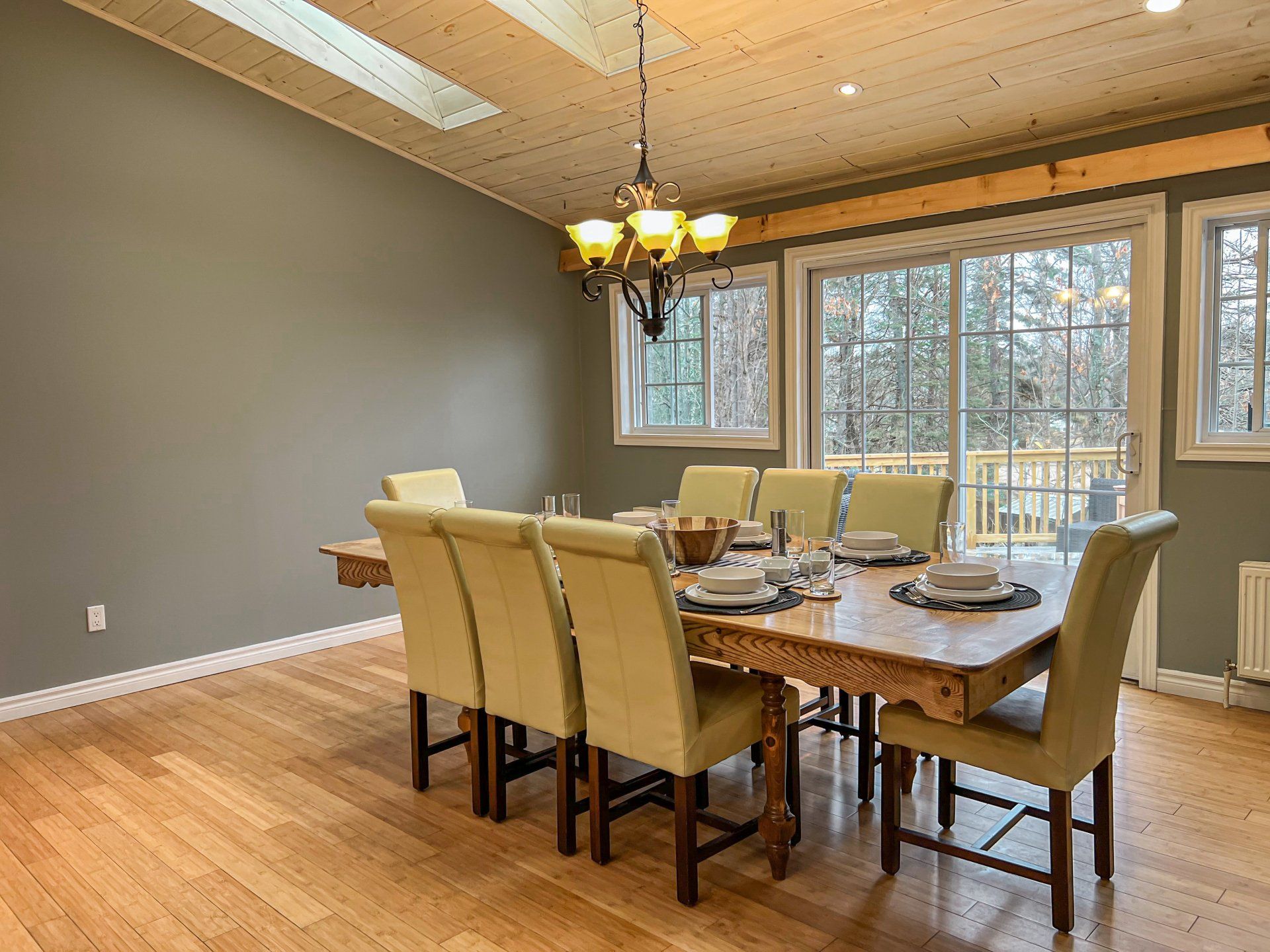 Dining room with wooden table, beige chairs, chandelier, and large windows overlooking trees.