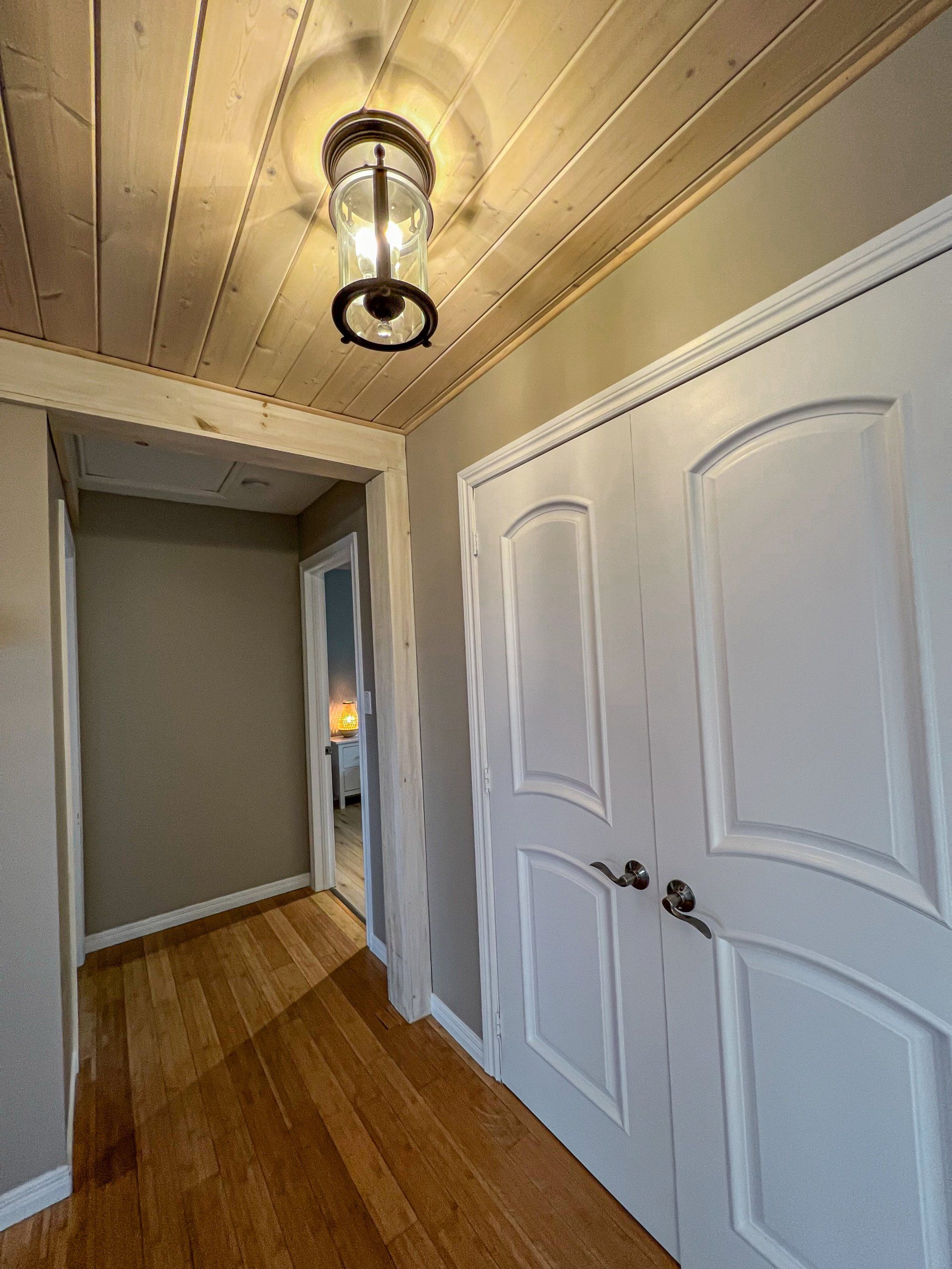 Narrow hallway with white double doors, wood floor, and ceiling light fixture; doorway glows warmly at the end.