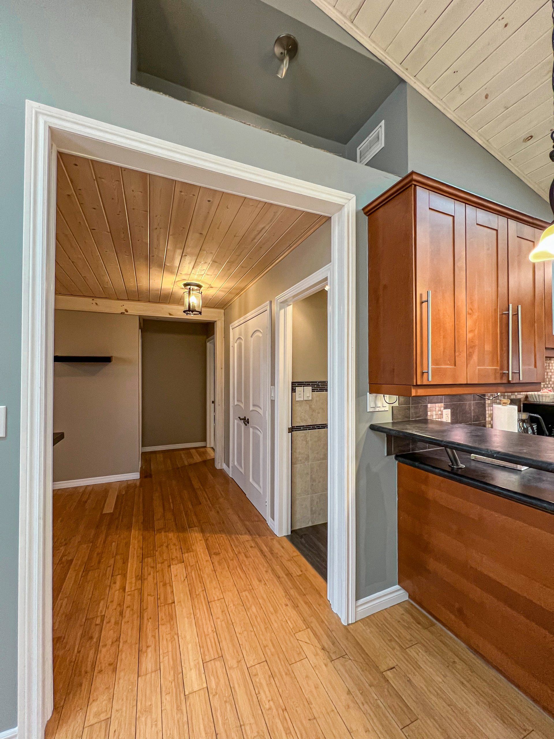 Kitchen opening to a narrow hallway with wood floors, gray walls, and wooden cabinets on the right.