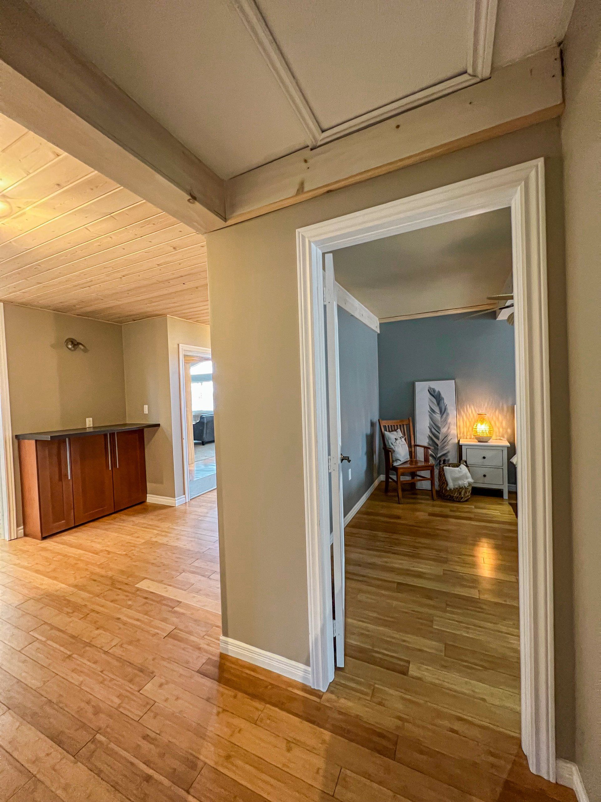 Hallway with hardwood floors, leading into a blue room with a lit lamp and chair.