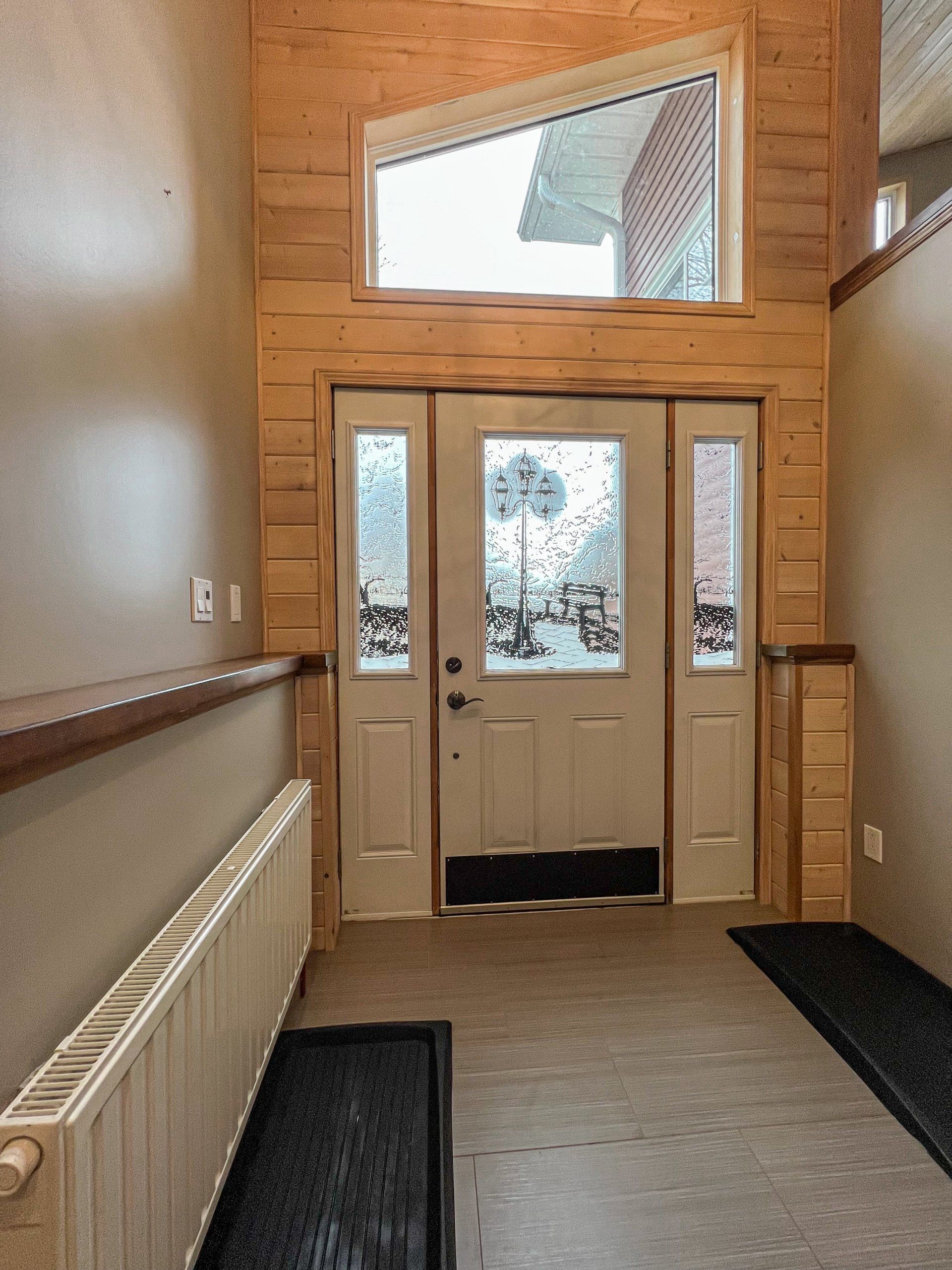 Front entry hallway with double glass doors, wood-paneled walls, and narrow black rugs on a gray floor.