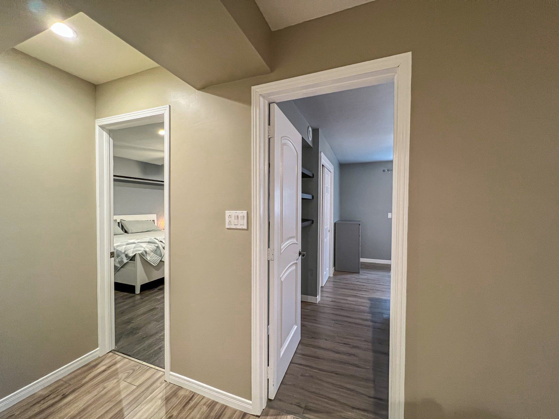 Hallway with beige walls, wood floors, and open doorways leading to a bedroom and another room.