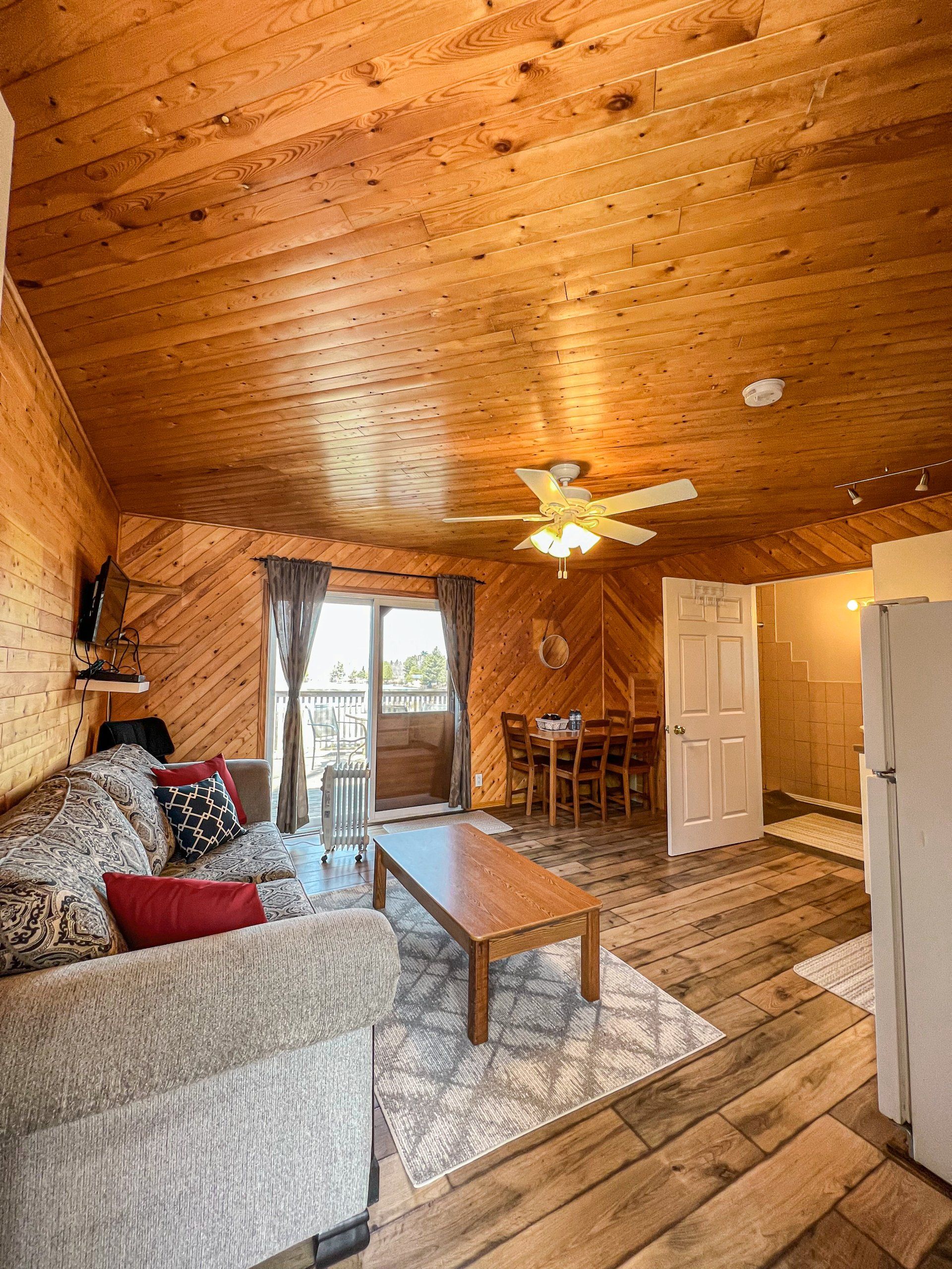 Cozy living room with wood-paneled ceiling, sofa, coffee table, rug, ceiling fan, and open door to a porch.