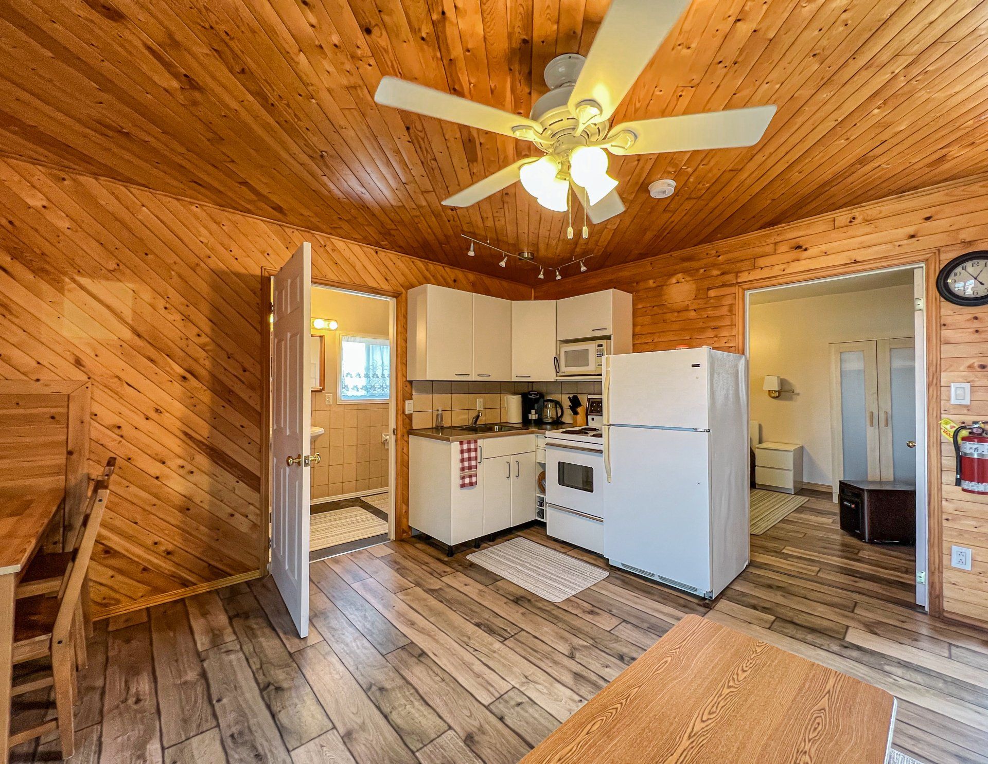 Cozy wood-paneled kitchen and living area with vaulted ceiling, ceiling fan, white appliances, and rustic floors