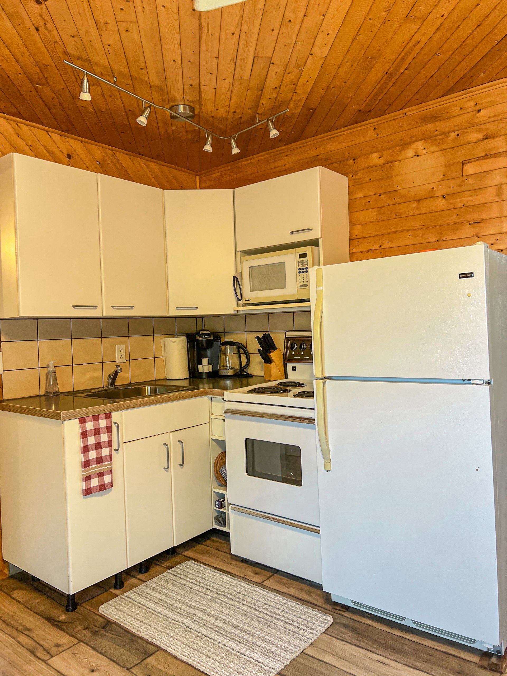 Small kitchen with white cabinets and appliances, wood-paneled ceiling, and a checkered towel on the counter.