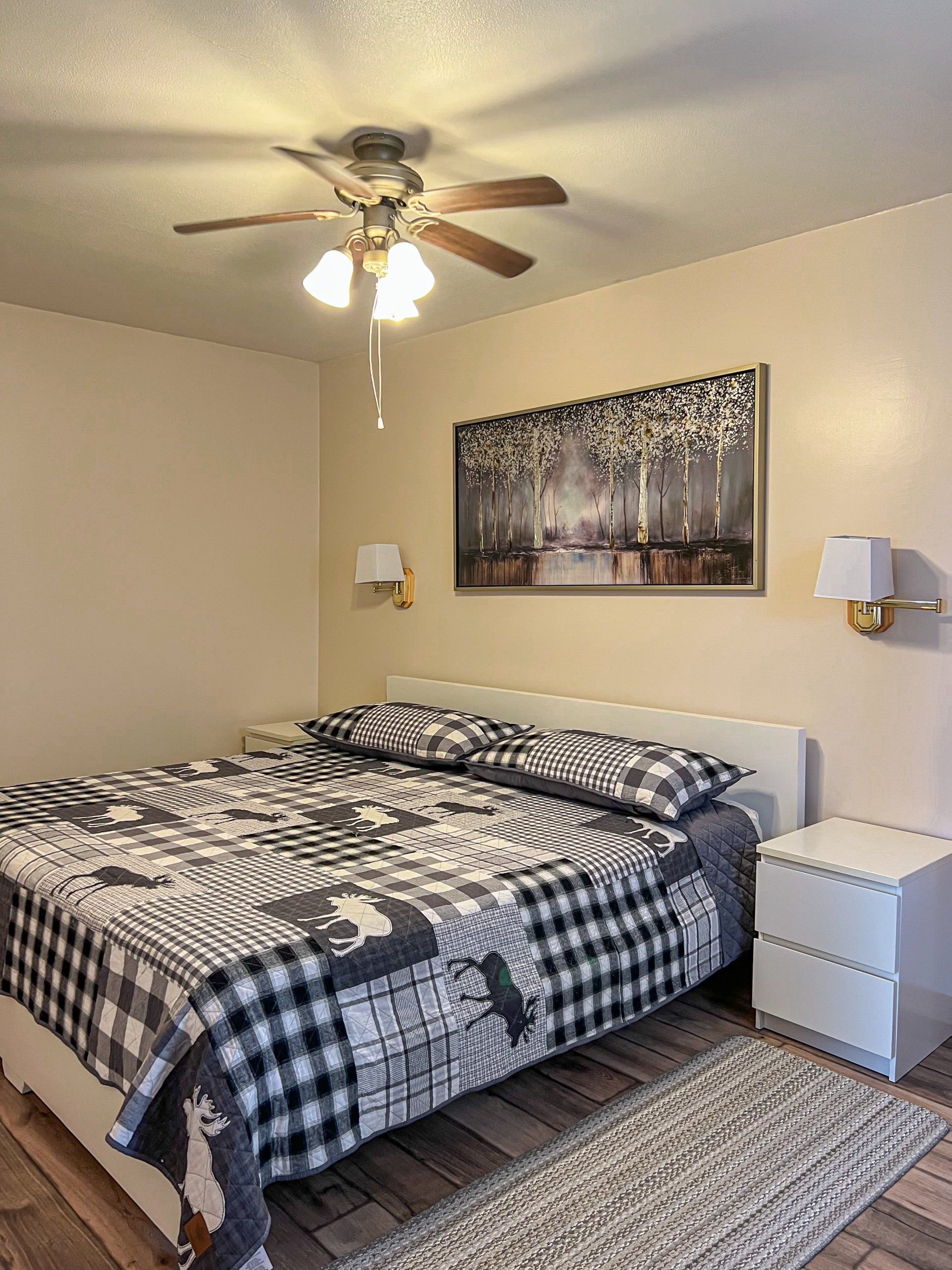 Bedroom with a neatly made black-and-white bed, ceiling fan, nightstands, and a forest wall print.