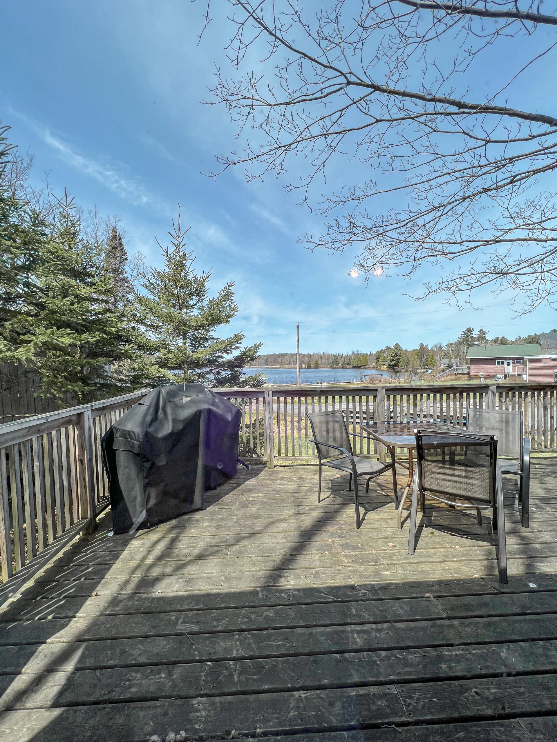 Sunny backyard deck with patio chairs, grill, and a view of trees and water under a blue sky
