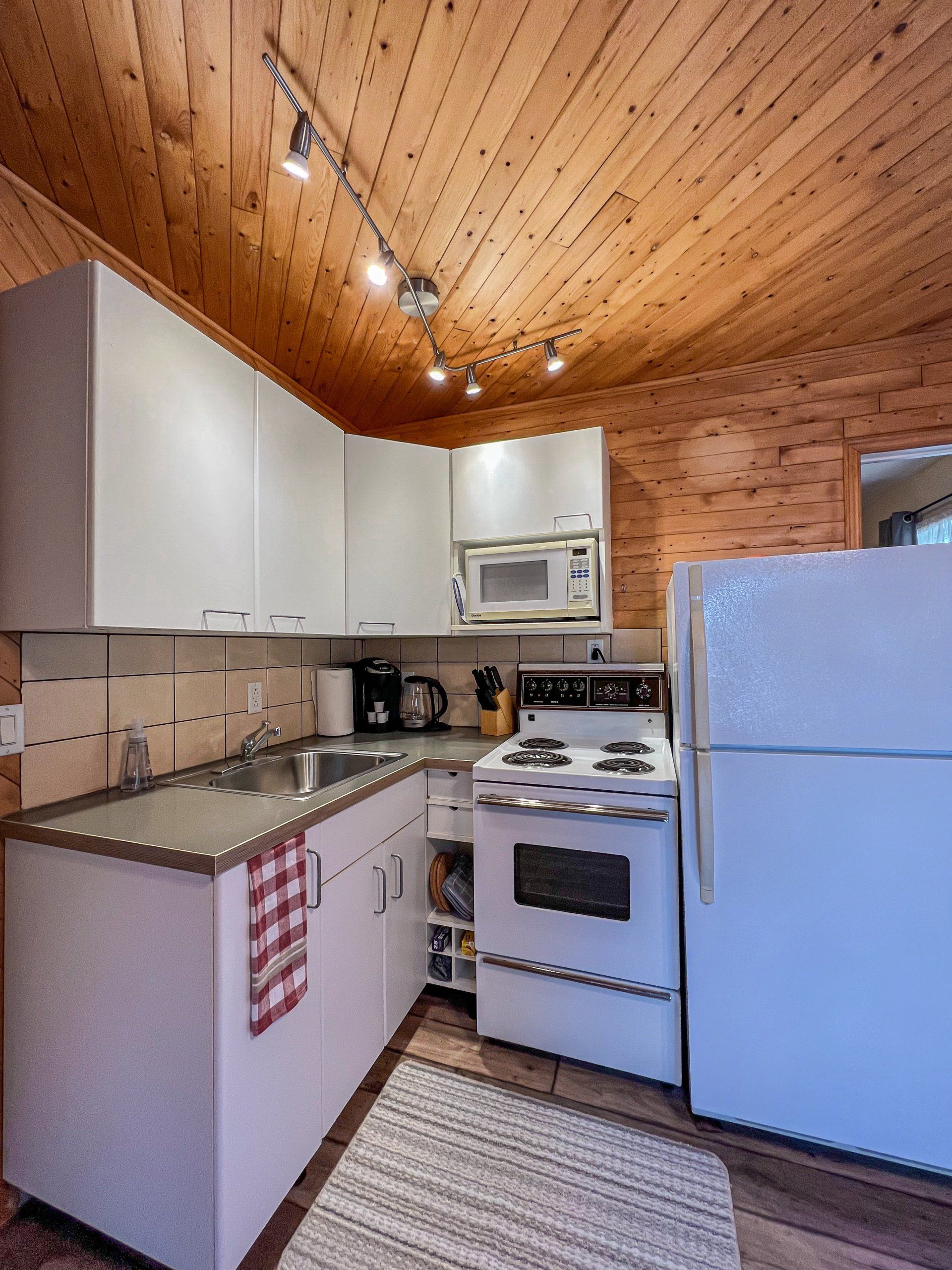 Compact kitchen with white appliances, wood-paneled ceiling, and light cabinets