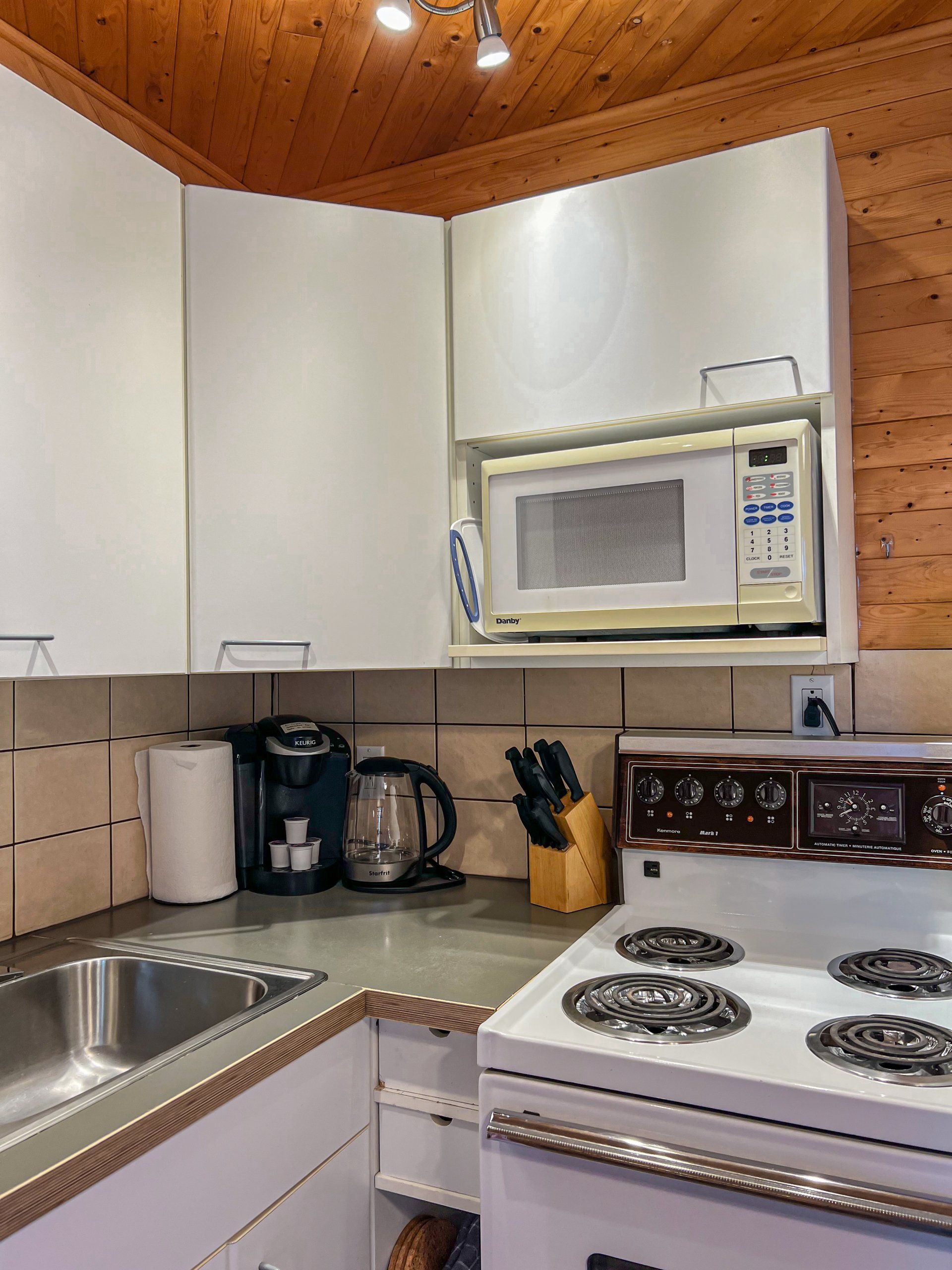 Small kitchen with white cabinets, stove, microwave, sink, coffee maker, and wood-paneled ceiling
