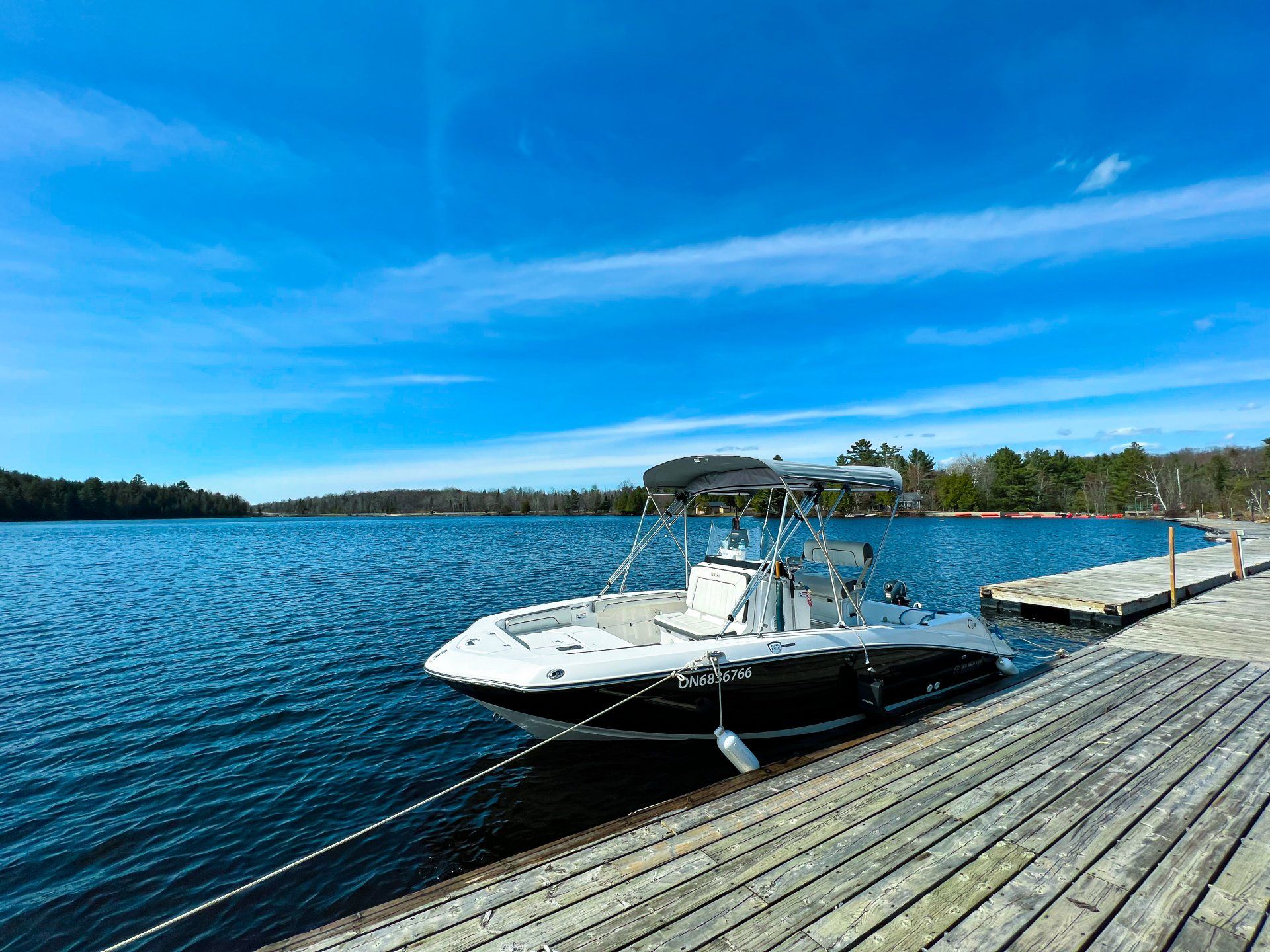 White motorboat docked beside a wooden pier on a sunny blue lake, with shoreline trees in the distance
