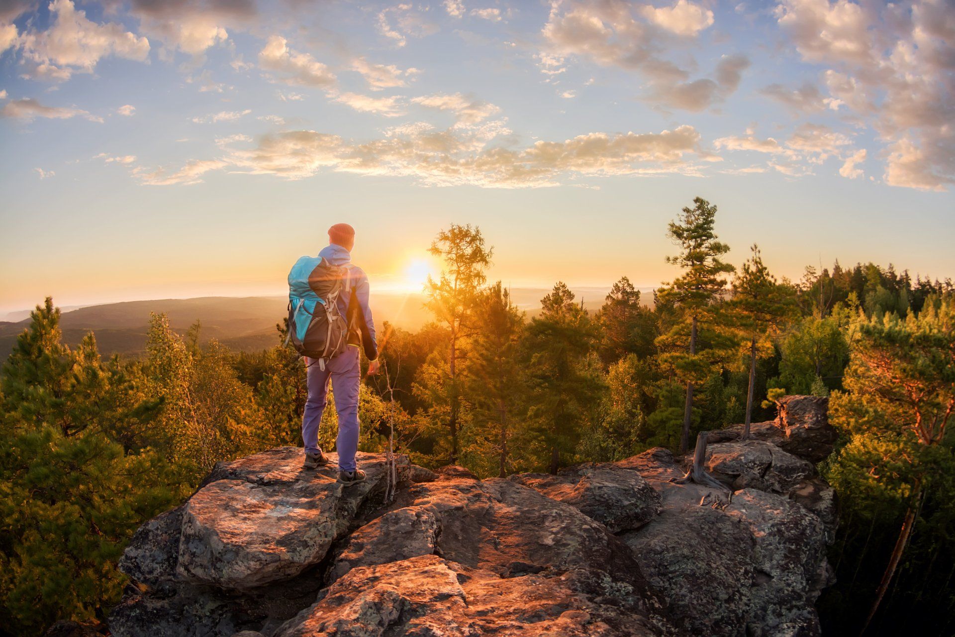Hiker with teal backpack stands on rocky cliff at sunset over a forested landscape.