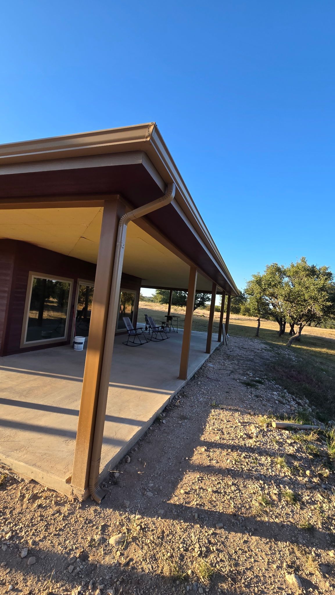 A wide patio with wooden support beams under a house roof, overlooking a sunny, gravel-filled yard with trees.