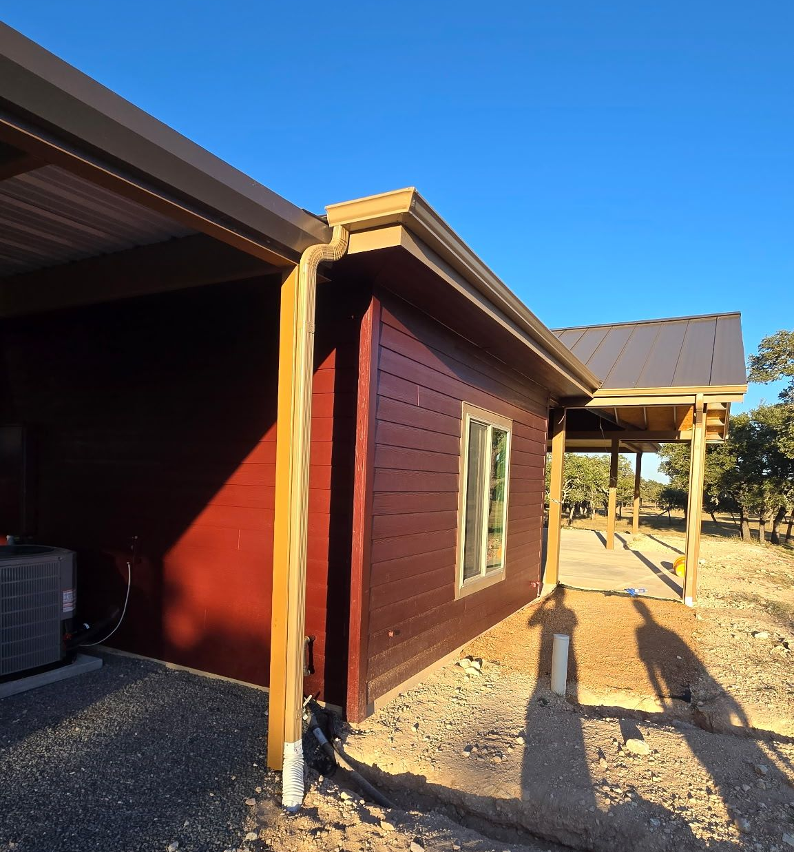 A dark red, horizontal-paneled house exterior with a metal roof, wooden support posts, and gravel ground at sunset.