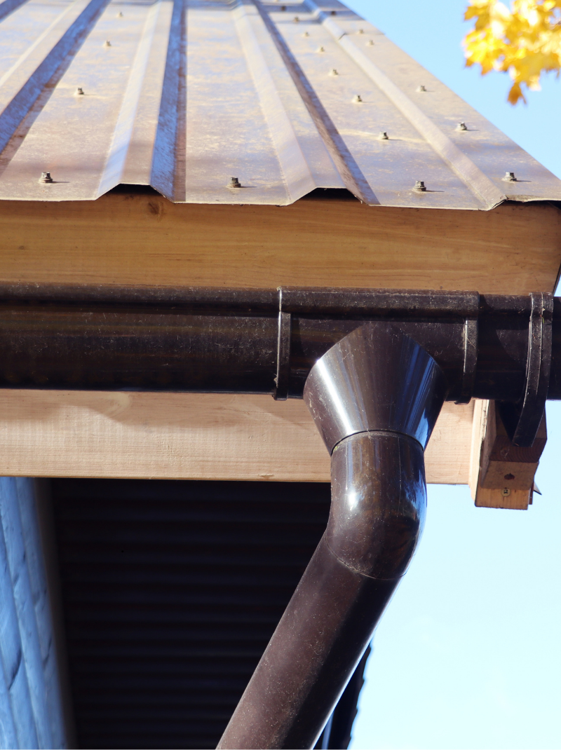 A close-up view of a metal roof edge with a brown gutter and downspout attached to a wooden fascia board.