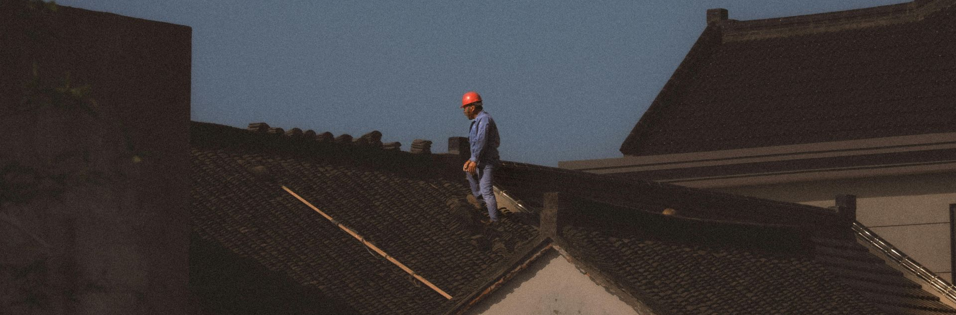 A person wearing a hard hat walks on the dark, sloped tiled roof of a traditional building.