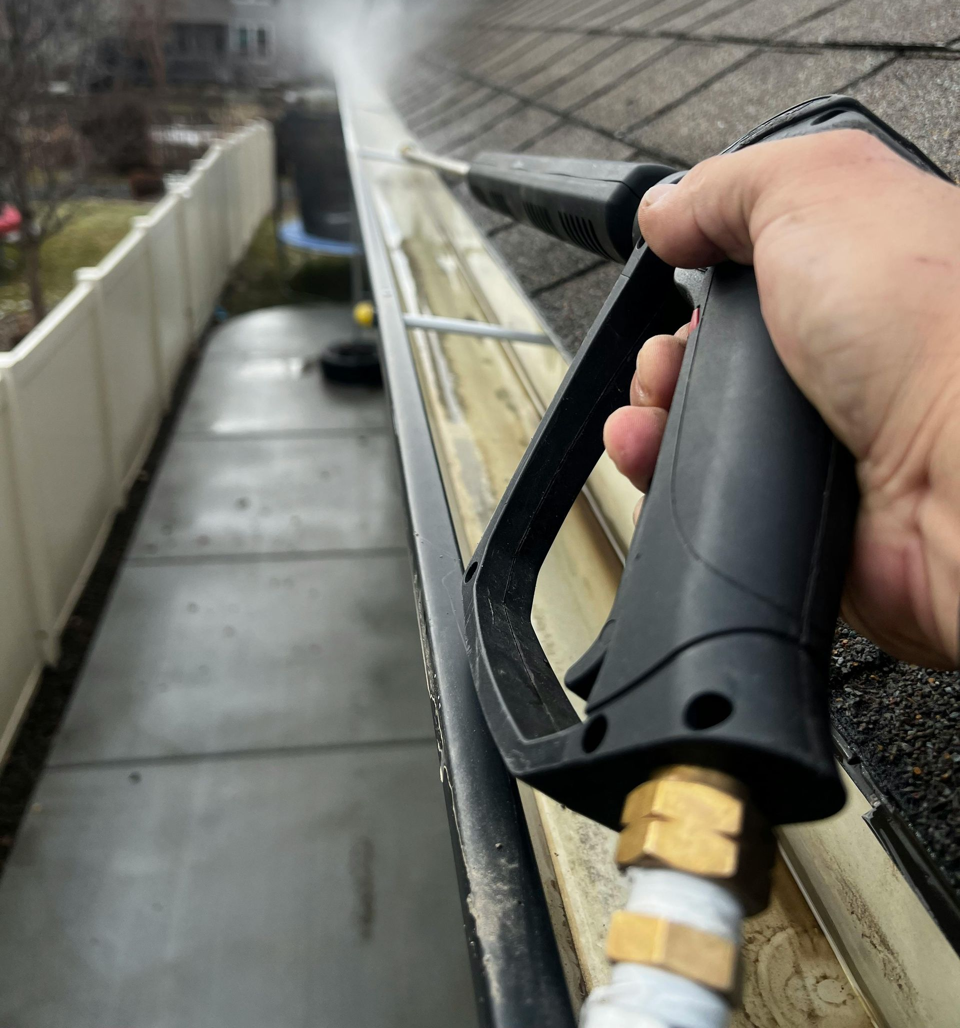 A hand holding a pressure washer wand sprays water to clean a residential gutter on the edge of a shingled roof.
