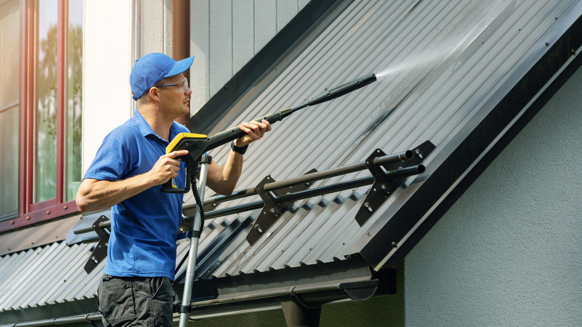 A worker in a blue shirt and cap uses a pressure washer to clean a dark, sloped metal roof.