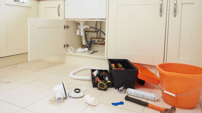Open cabinet under a sink with plumbing tools, hoses, and buckets on a kitchen floor