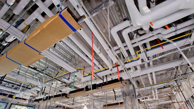Industrial ceiling with exposed pipes, ducts, and wiring, including red hanging straps and cardboard-covered sections