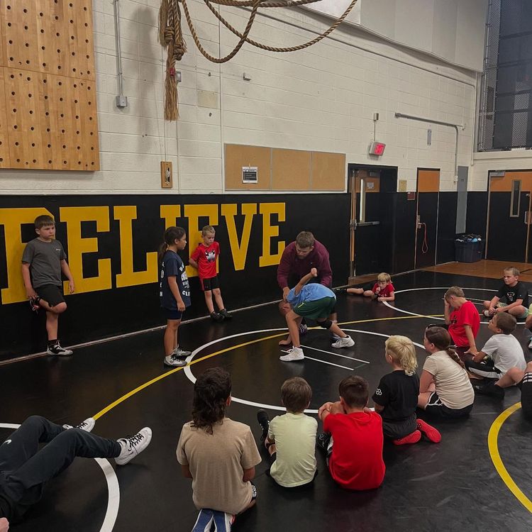 Gym class with students and instructor on a wrestling mat in a gymnasium with “ELITE” on the wall