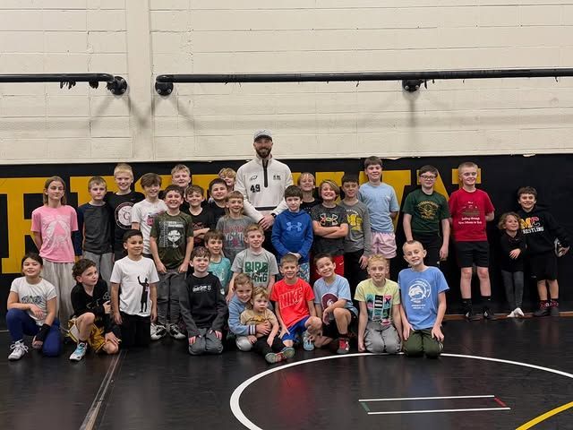Large group posing in a gym on wrestling mats, with a coach standing behind them.