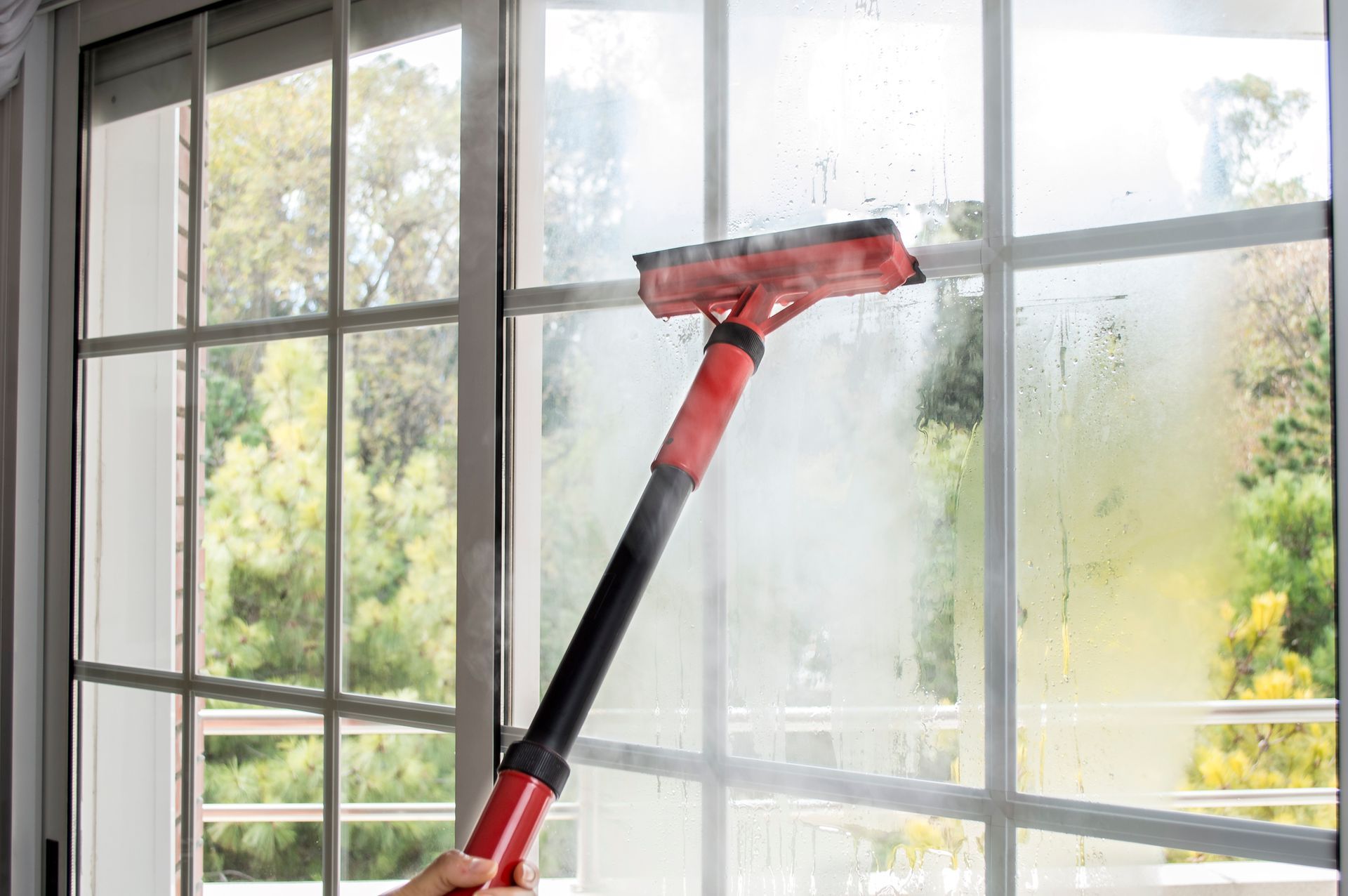 A person using a red squeegee to clean a window with condensation, set against trees.