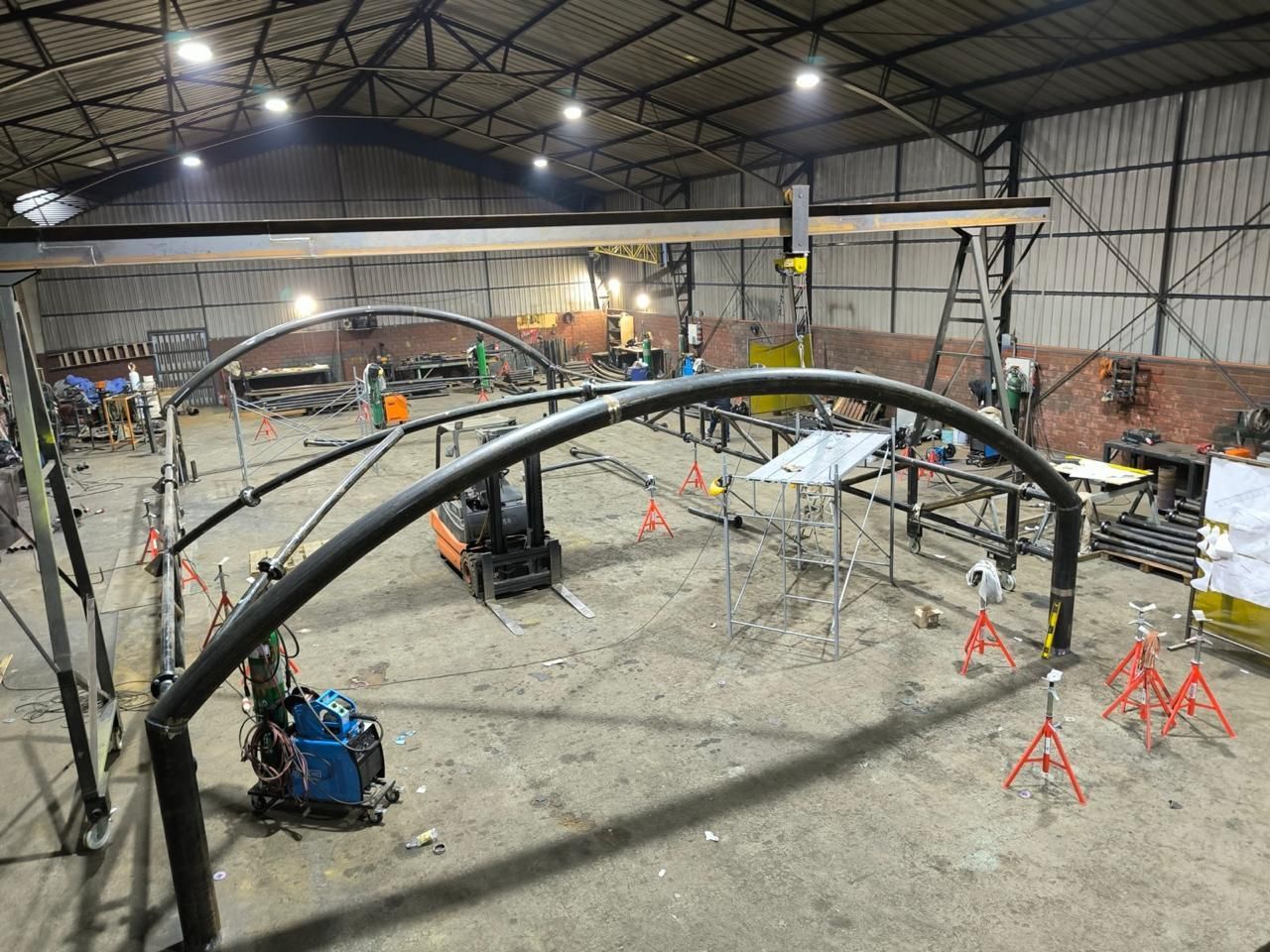 Interior de fábrica con estructuras metálicas arqueadas en construcción, carretilla elevadora presente.