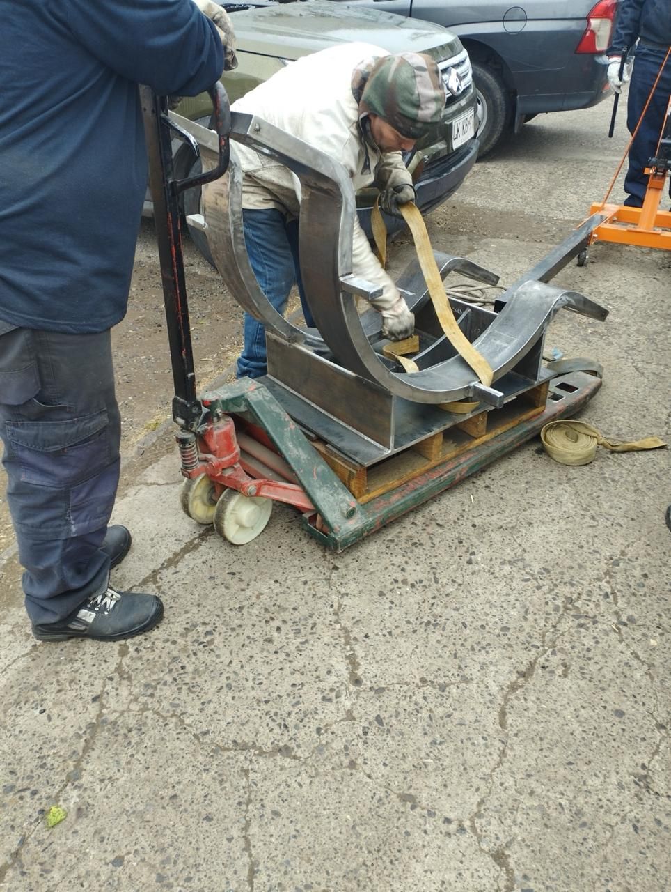 Un hombre usa una transpaleta para mover un objeto metálico grande sobre un palé. Al aire libre, cerca de un coche.