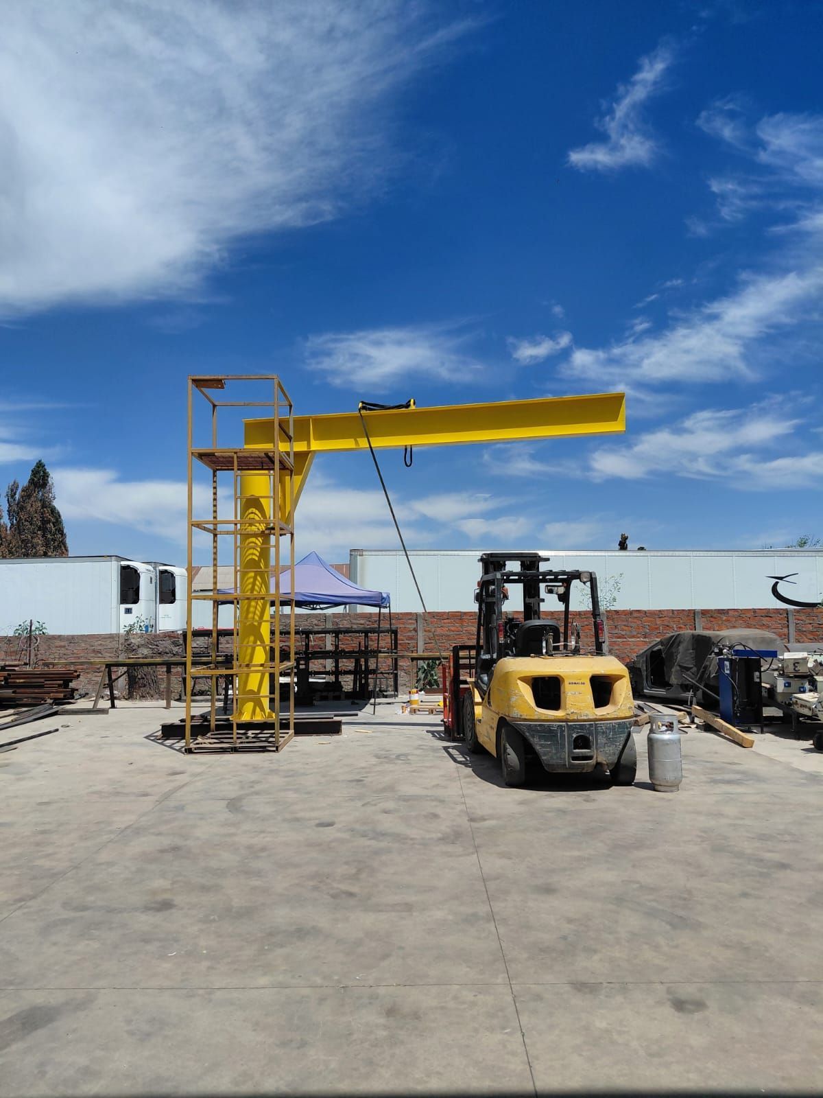 Grúa pluma amarilla con carretilla elevadora en un entorno industrial al aire libre.