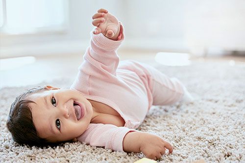 Smiling baby in a pink onesie lying on a fluffy rug in bright daylight