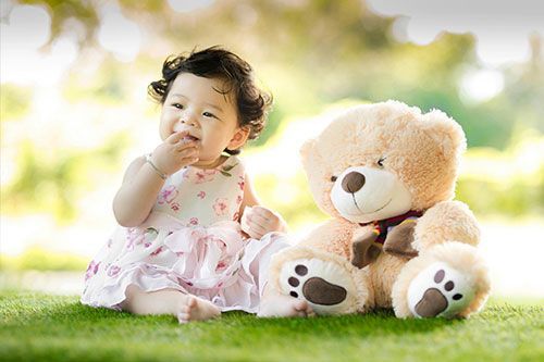 Baby in a pink dress sitting on grass beside a teddy bear in a sunlit park