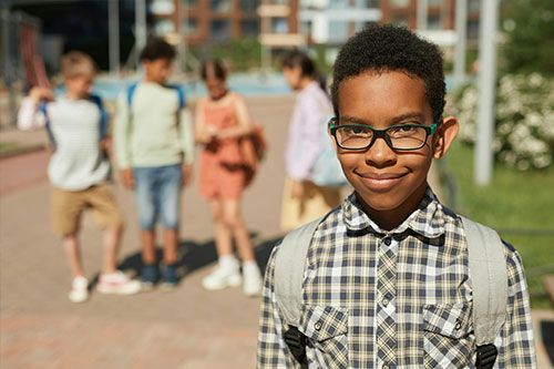 Smiling child in glasses and plaid shirt standing outdoors with other children blurred in the background
