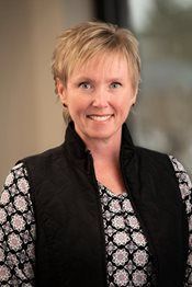 Smiling woman in black vest and patterned blouse against a blurred indoor background