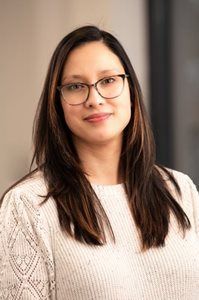 Portrait of a person with glasses and long dark hair, wearing a white sweater against a neutral background