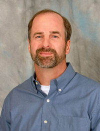Portrait of a man with a beard wearing a blue button-down shirt against a gray backdrop