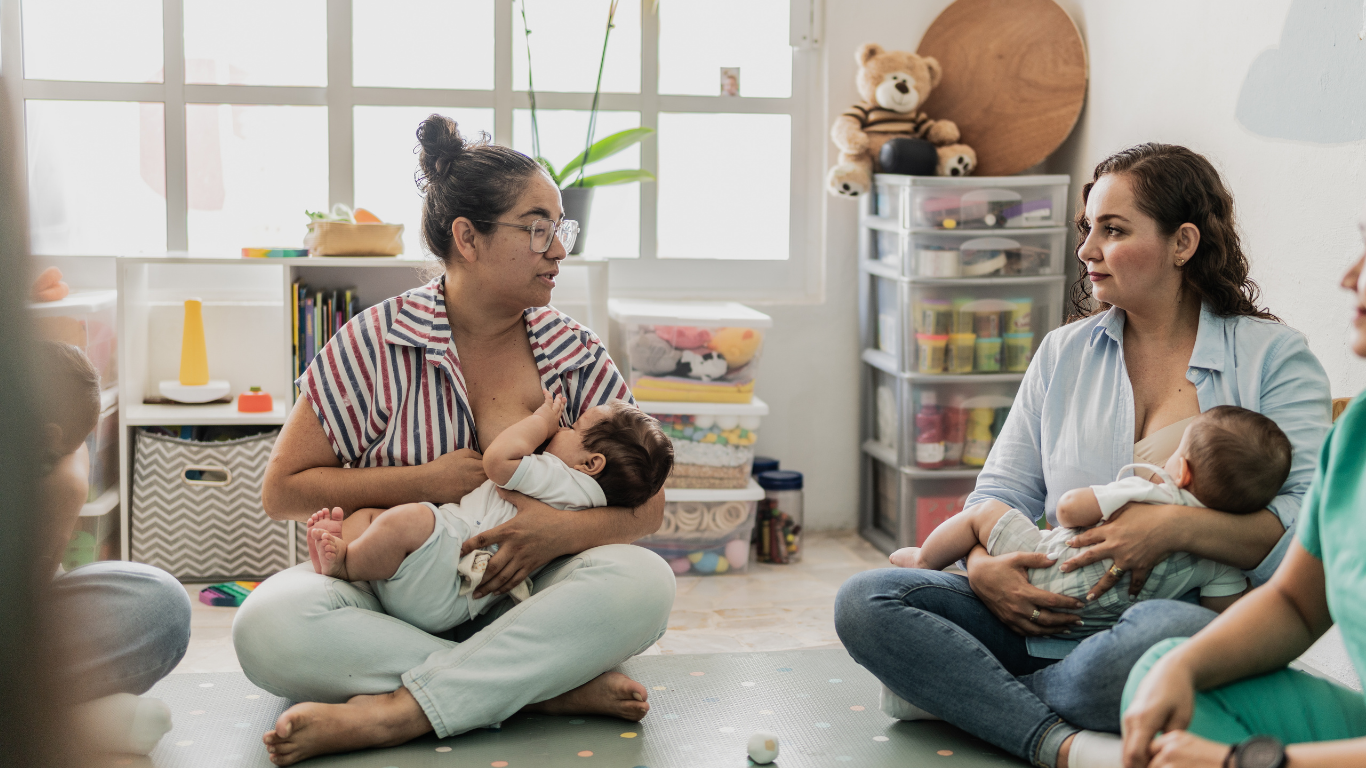 Click to Visit Calendar Two caregivers feeding infants in a bright playroom, seated on the floor with toys and shelves behind them