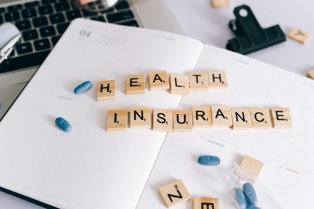 Scrabble tiles spelling “HEALTH INSURANCE” on papers with blue pills and a laptop keyboard nearby