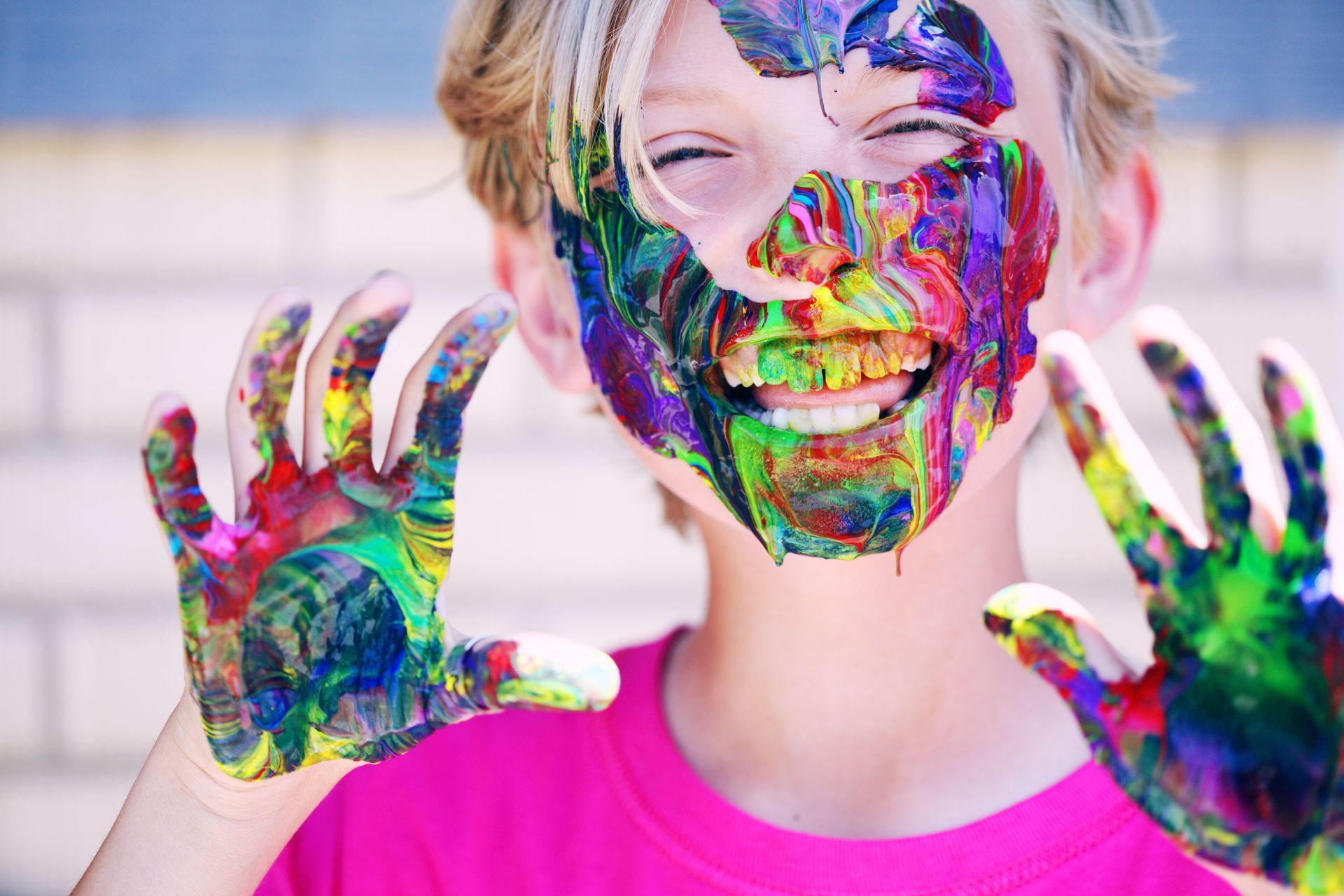 Child smiling with multicolored paint on face and hands, wearing a bright pink shirt