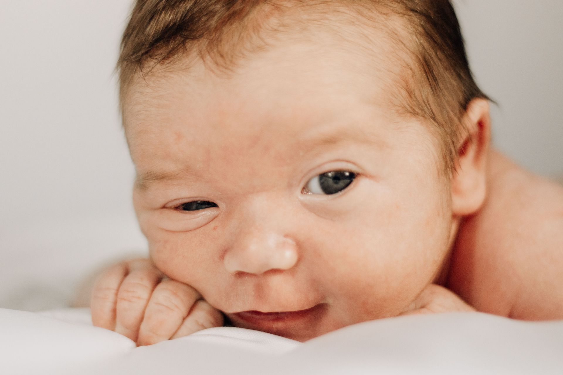 Close-up of a newborn lying on a white blanket, resting on hands and looking at the camera