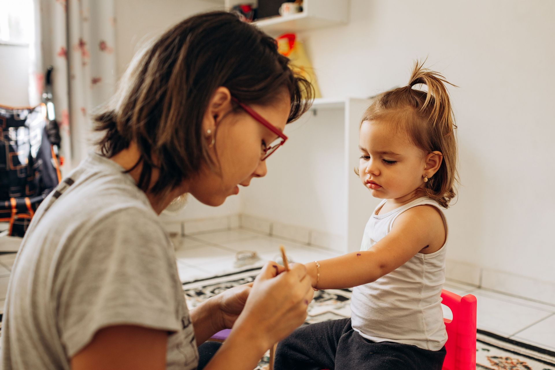 Adult painting a child’s arm in a bright kitchen, both focused on the activity.
