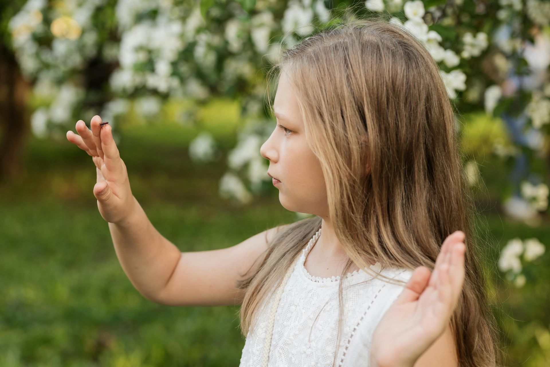 Young child in a white dress reaching out with both hands in a flower-filled garden