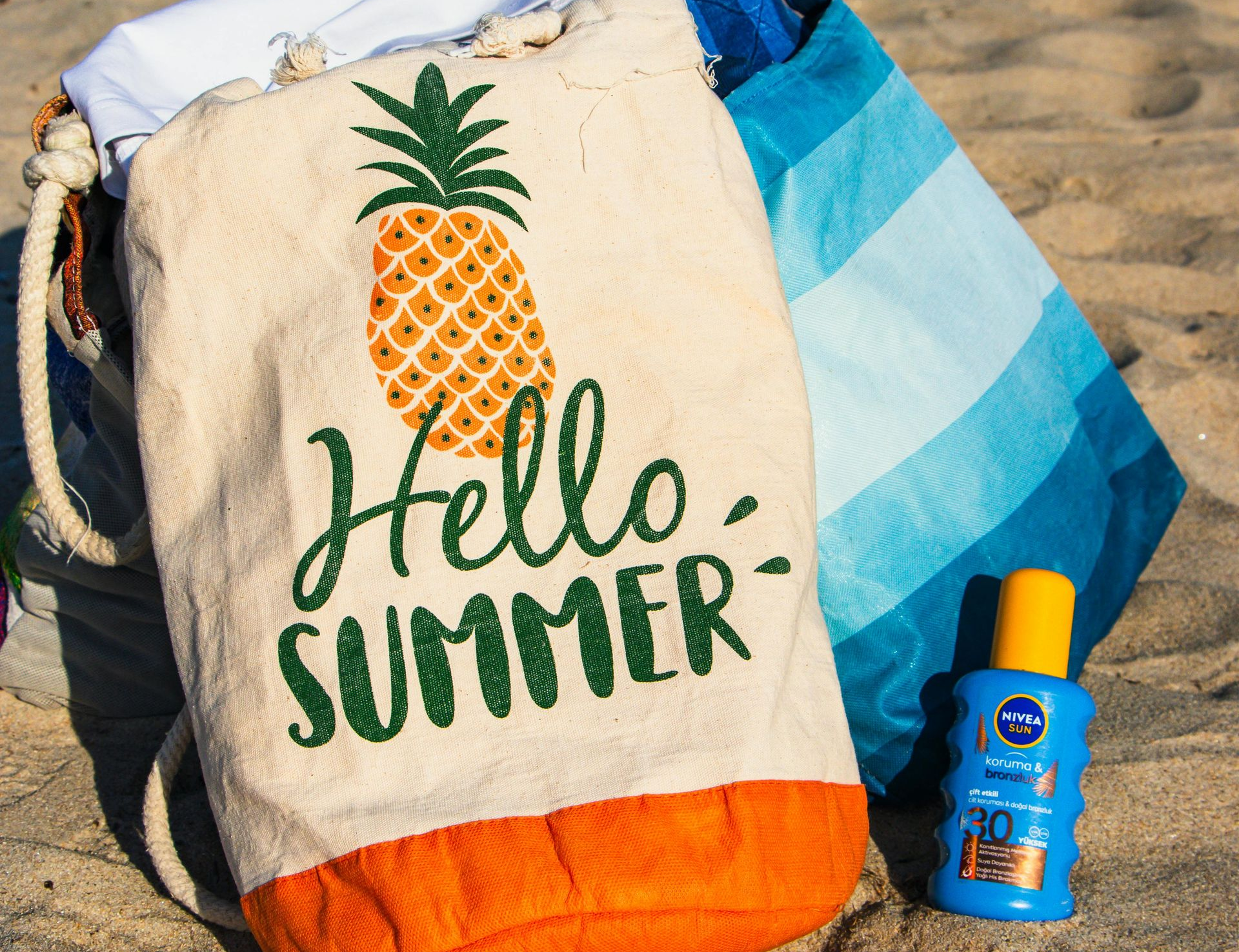 Beach bag with pineapple and “Hello Summer” text beside sunscreen on sand
