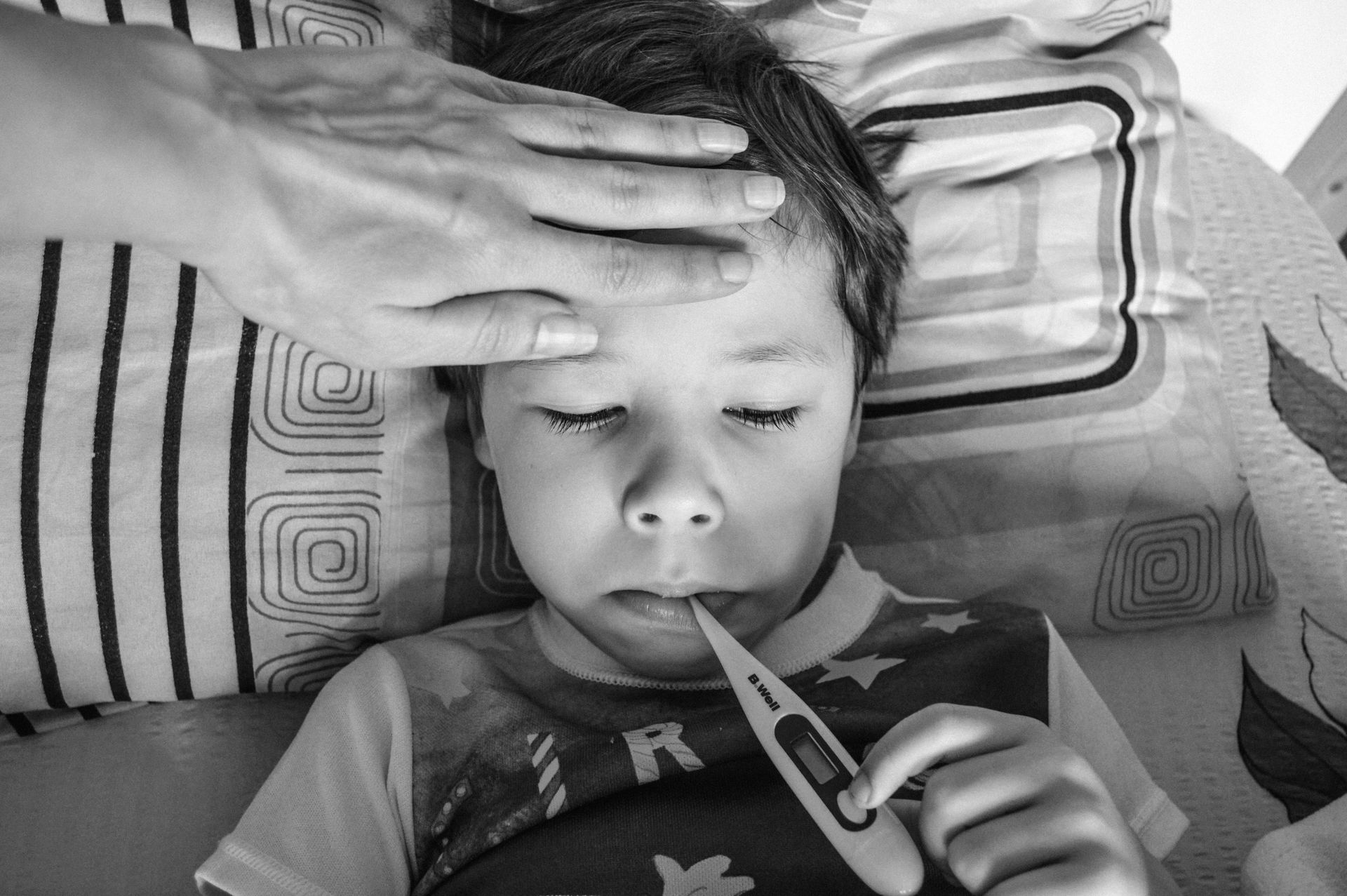 Child lying in bed with a thermometer, while an adult hand rests on their forehead