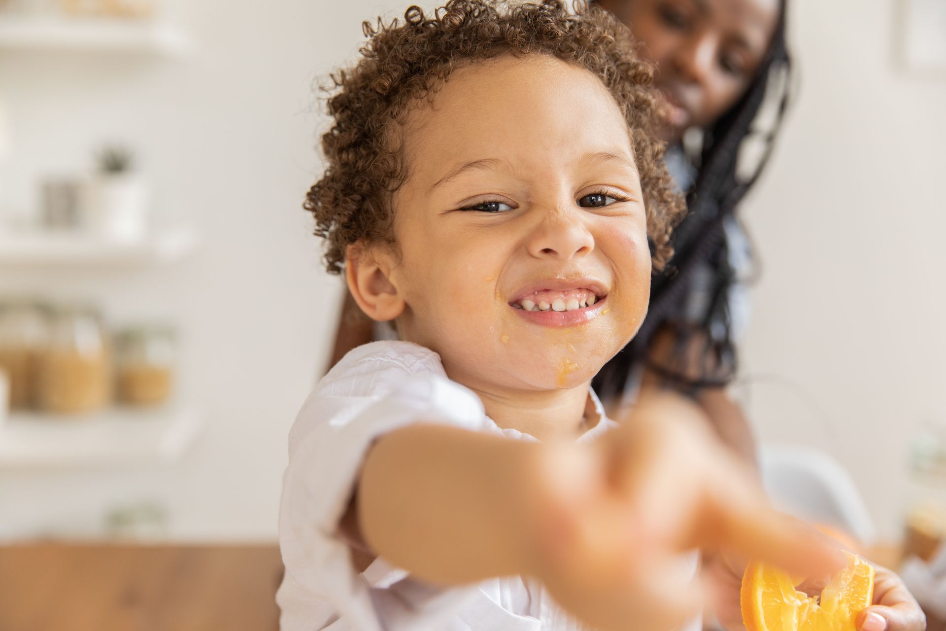 Smiling child reaching toward the camera in a bright kitchen, with an adult in the background