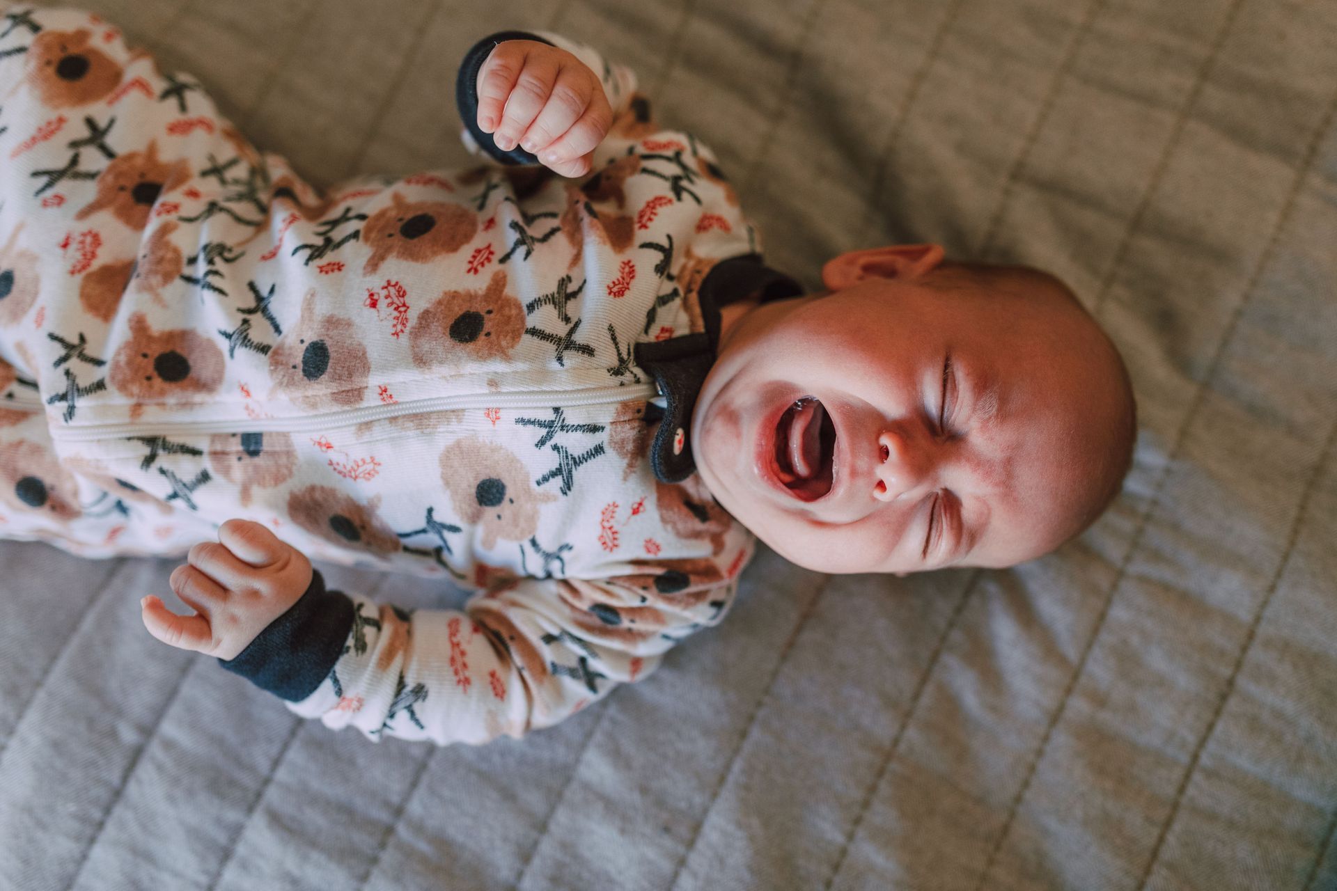 Crying baby in a patterned onesie lying on a gray quilted blanket