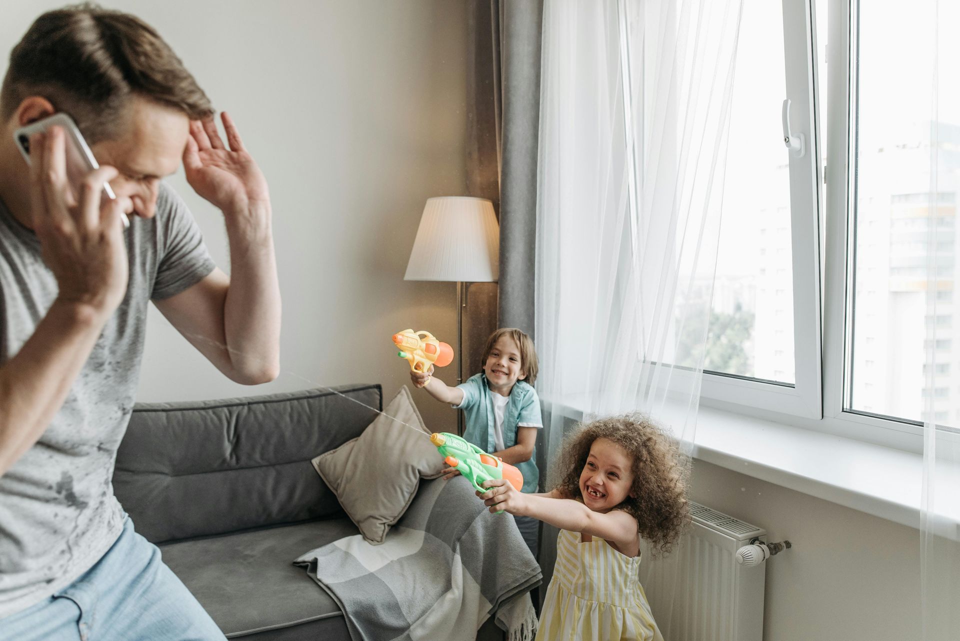 Adult on phone in a living room while two children play near a window.