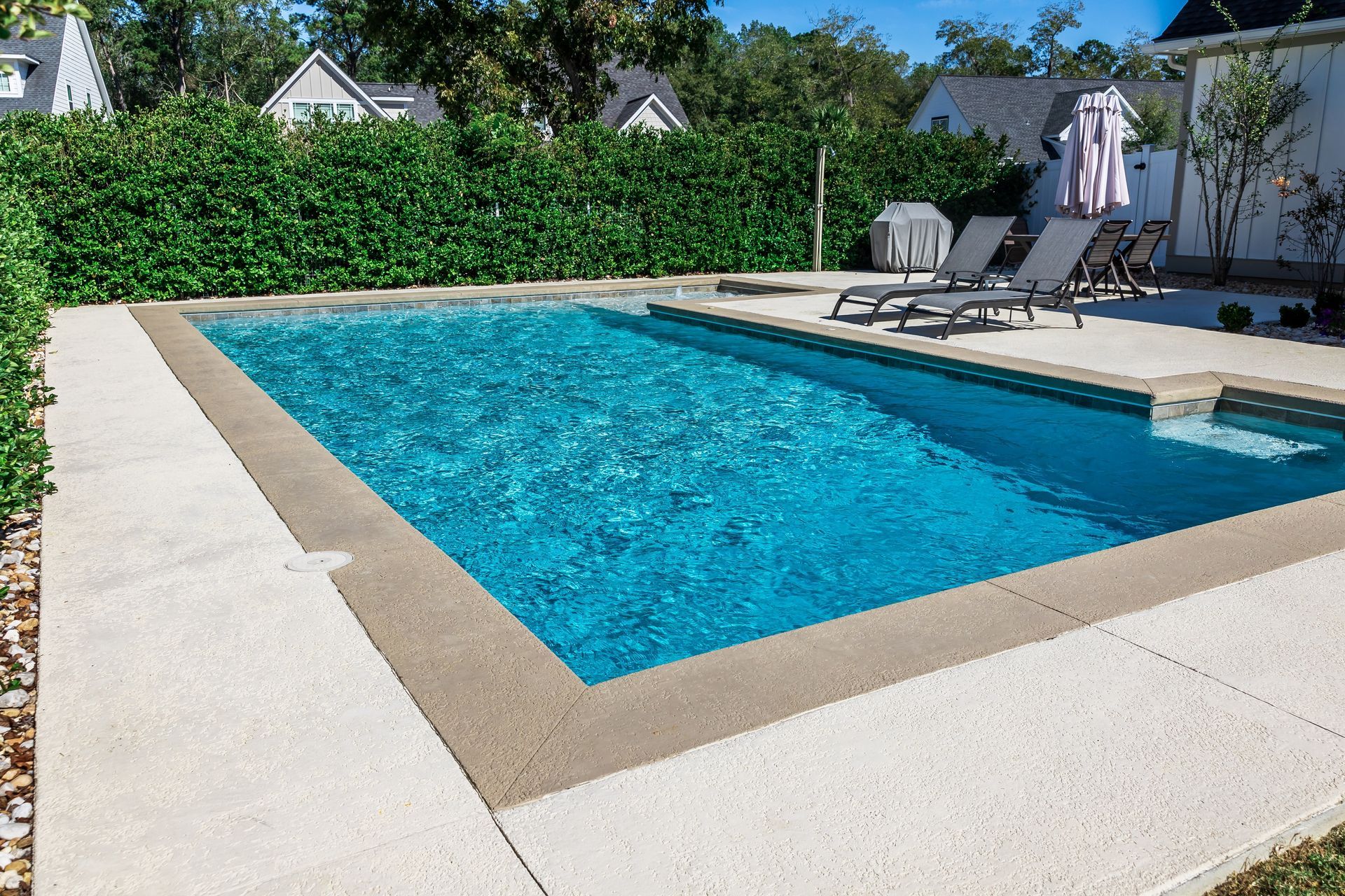 Outdoor swimming pool with blue water, beige deck, lounge chairs, and greenery in the background